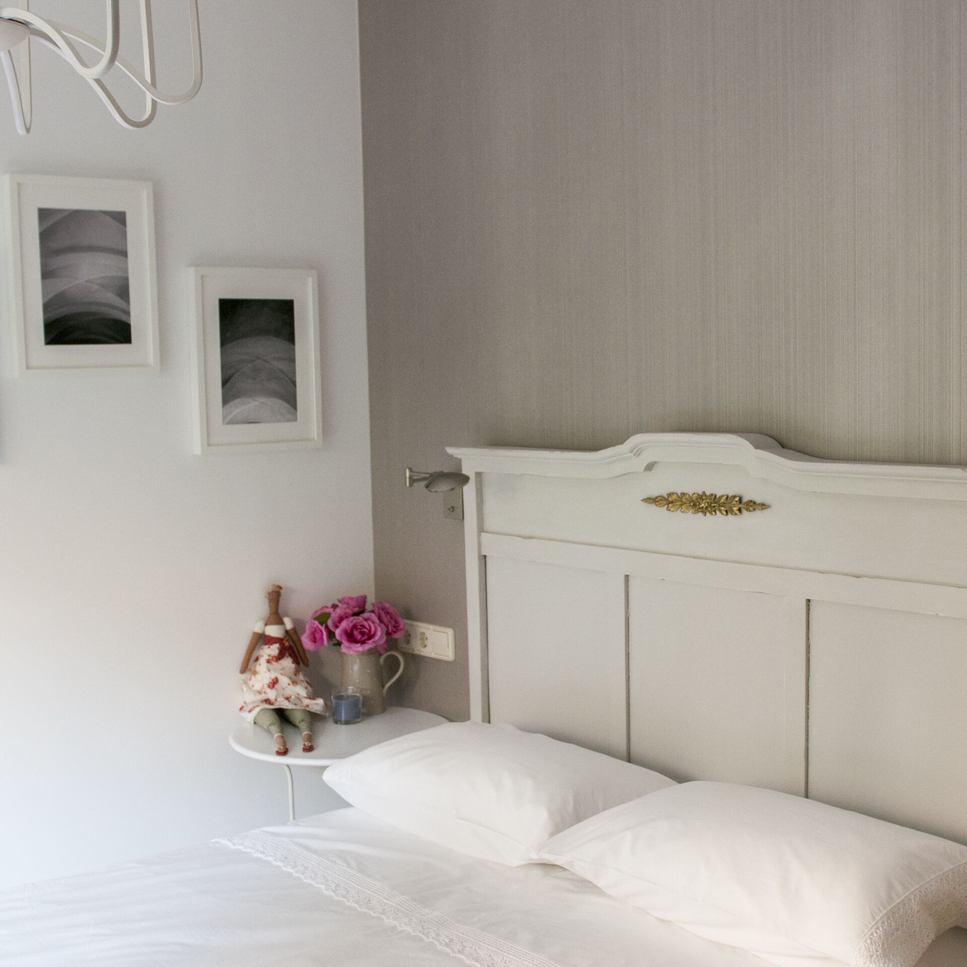 Um quarto com uma cama branca, uma mesa de cabeceira com um vaso de flores e duas fotografias emolduradas na parede.
