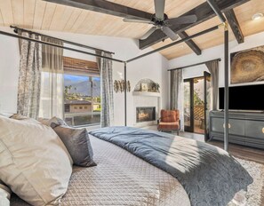 A bedroom with a large bed, a ceiling fan, a fireplace, and a view of the outside.