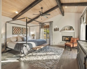 A four-poster bed with a bench, a fireplace, and a ceiling fan in a room with wooden beams and a large window.