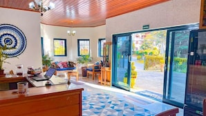 A hotel lobby with a wooden ceiling, a blue and white patterned wall, a reception desk, a sofa, a coffee table, and a large glass door leading outside. - Castelo Piazzale by Concavus (Campos do Jordão)