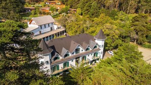 A large, multi-story house with a prominent tower and steep roof, surrounded by dense forest. - Castelo Piazzale by Concavus (Campos do Jordão)