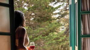 A woman in a long dress standing by a window with a view of trees, holding a glass of red wine. - Castelo Piazzale by Concavus (Campos do Jordão)