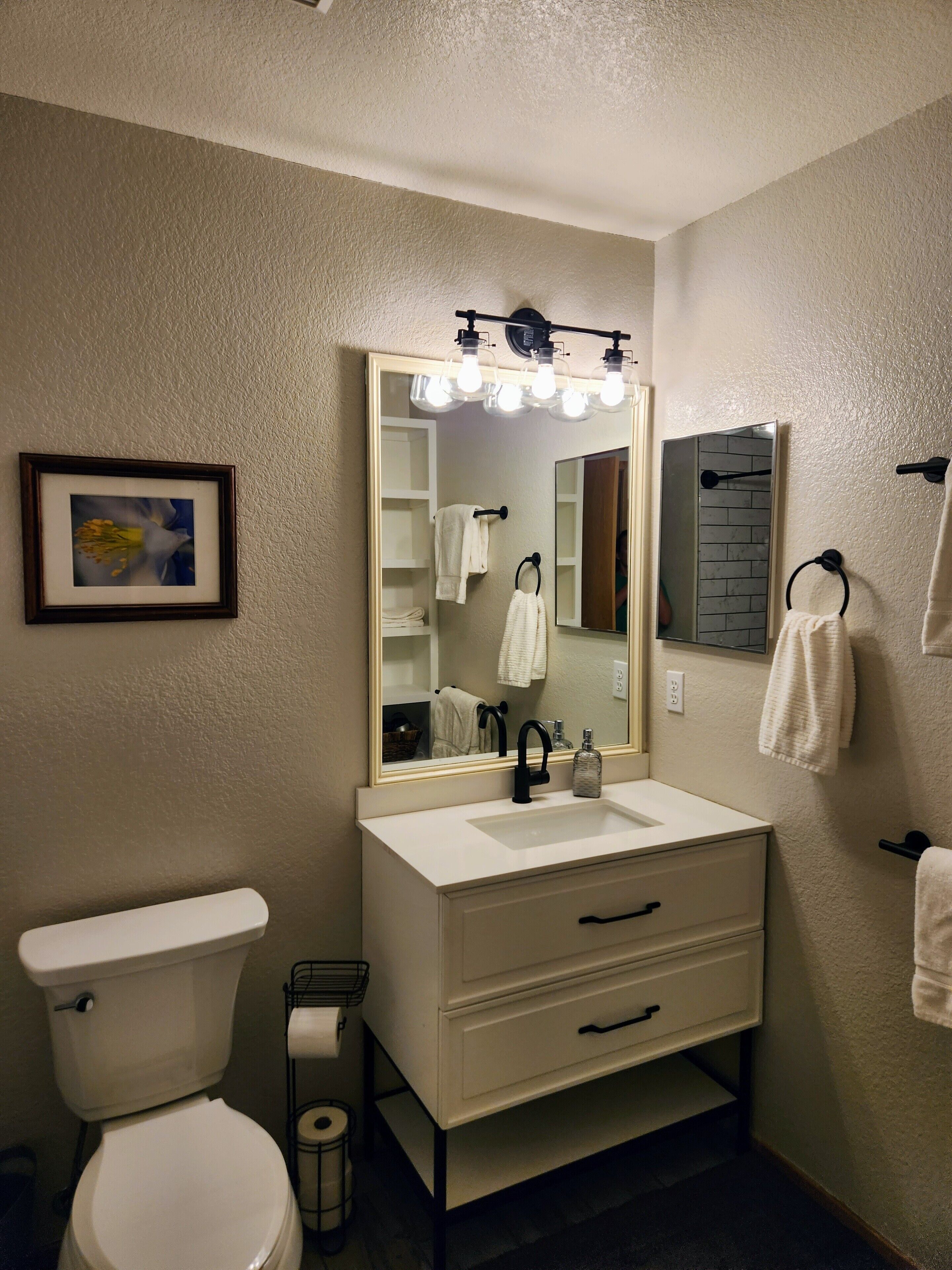 Une salle de bain avec un lavabo blanc, un miroir avec éclairage intégré, des toilettes et un tableau encadré accroché au mur.