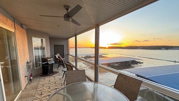 A balcony with a glass table, chairs, and a ceiling fan. A view of the sunset over a body of water is visible through the glass. - Lands End Condo 3Bd/3Bath End Unit with Amazing Main Channel View, Newly Updated (Osage Beach)