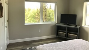 A bedroom with a large window, a bed, a TV on a stand, and a small shelf with baskets. - Cozy 1 bedroom with beautiful Alaskan views (Anchorage)