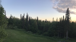 A view from a wooden deck overlooking a grassy slope with a forested area and a small outdoor seating area with a fire pit. - Cozy 1 bedroom with beautiful Alaskan views (Anchorage)