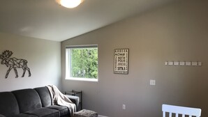 A living room with a gray couch, a small ottoman, a white chair, a rug, a window with a view of trees, and a wall with a moose artwork and a word art piece. - Cozy 1 bedroom with beautiful Alaskan views (Anchorage)