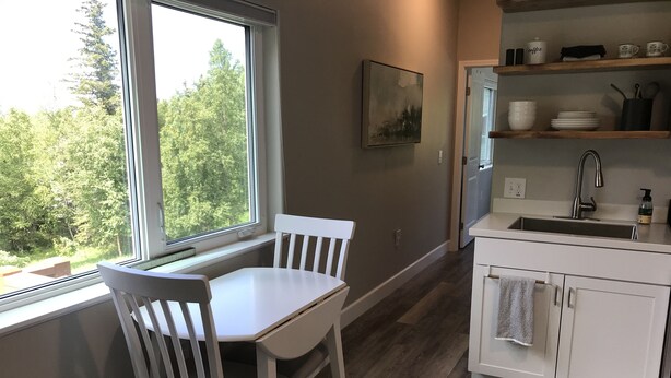 A kitchen with white cabinetry, a wooden floor, and a window overlooking greenery. - Cozy 1 bedroom with beautiful Alaskan views (Anchorage)