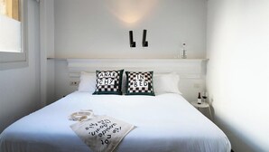 A neatly made bed with two decorative pillows, a bedside table with a bottle and glass, and a wall-mounted light fixture. - Basic Hotel (Olerdola)