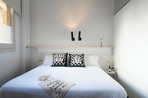 Una cama bien hecha con dos almohadones decorativos, una mesita de noche con una botella y un vaso, y una lámpara de pared.
