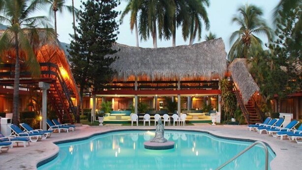 A pool area with lounge chairs, a central fountain, and a building with a thatched roof. - Bali Hai (Acapulco)