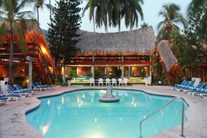 A pool area with lounge chairs, a central fountain, and a building with a thatched roof. - Bali Hai (Acapulco)