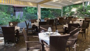 Un restaurante al aire libre con sillas y mesas de mimbre, cubierto por un techo y rodeado de follaje.