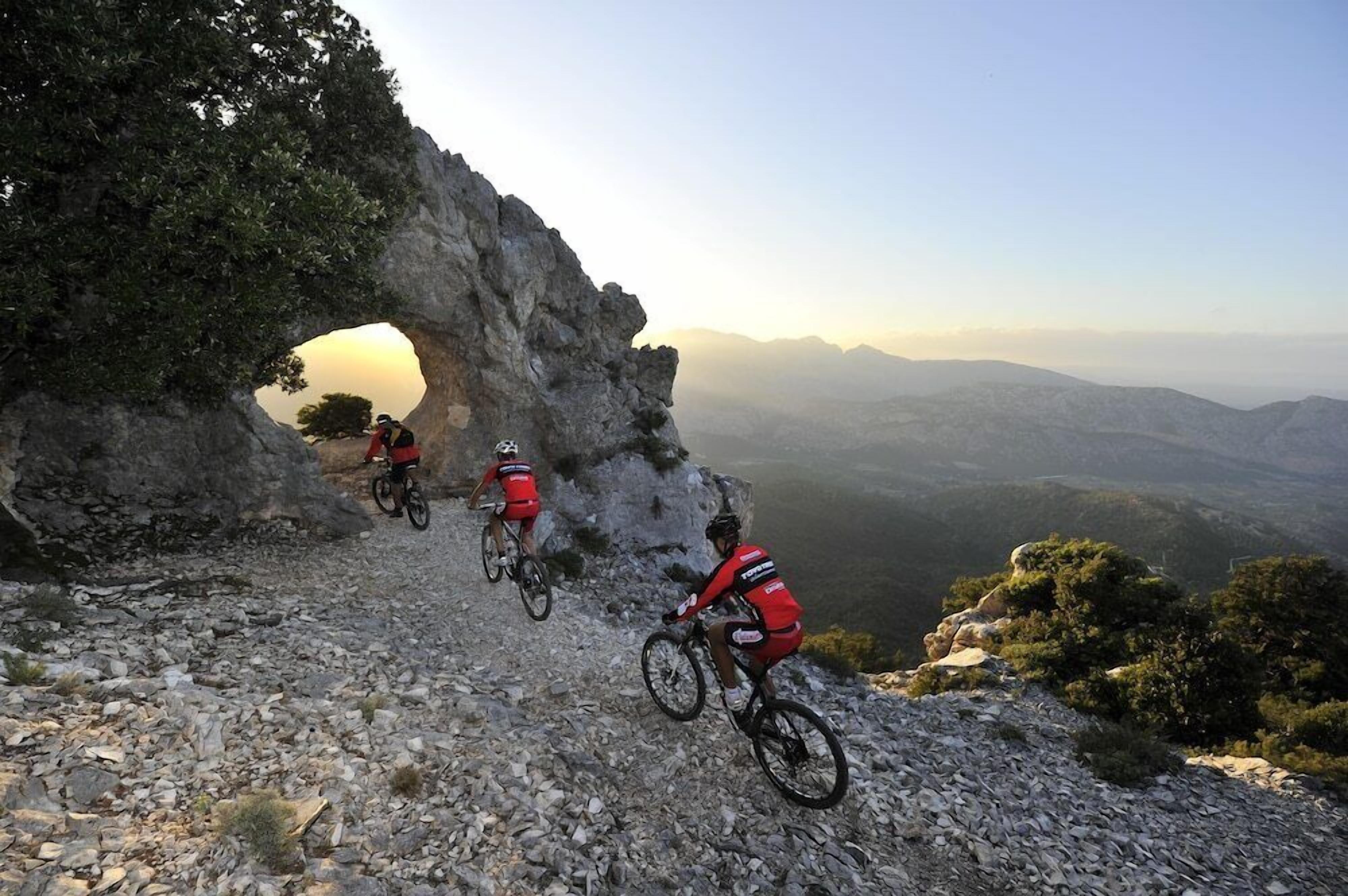 Three cyclists on a rocky mountain trail with a scenic view of distant mountains and a clear sky.