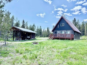 A rustic cabin with a covered porch and a large wooden deck, surrounded by a grassy field and dense forest. - Holden Hideaway - 23 miles to YNP ~ Outdoor kitchen ~ Firepit ~ Wifi (Island Park)