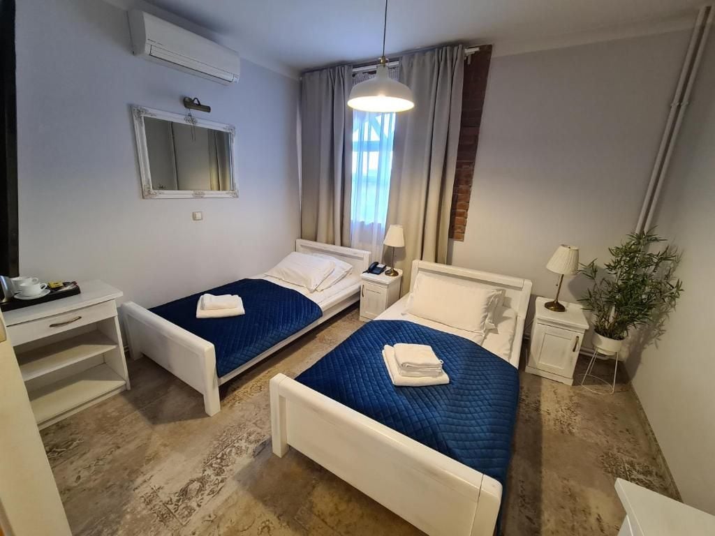 A hotel room with two single beds, a desk, a chair, a small table, a lamp, and a potted plant.