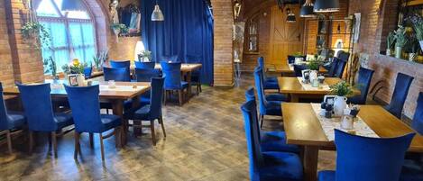 A restaurant with brick walls, wooden beams, and blue upholstered chairs. Tables are set with plates, cups, and cutlery.