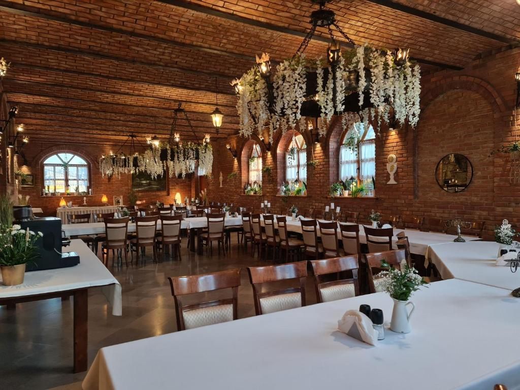 A spacious dining hall with brick walls, arched windows, and hanging floral decorations. Tables are set with white tablecloths and chairs.