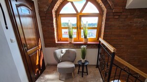 A room with a wooden door, a grey armchair, a small table with a plant, a yellow pendant light, and a window with a view of a building. - Hotel Staropolski (Tomaszow Lubelski)