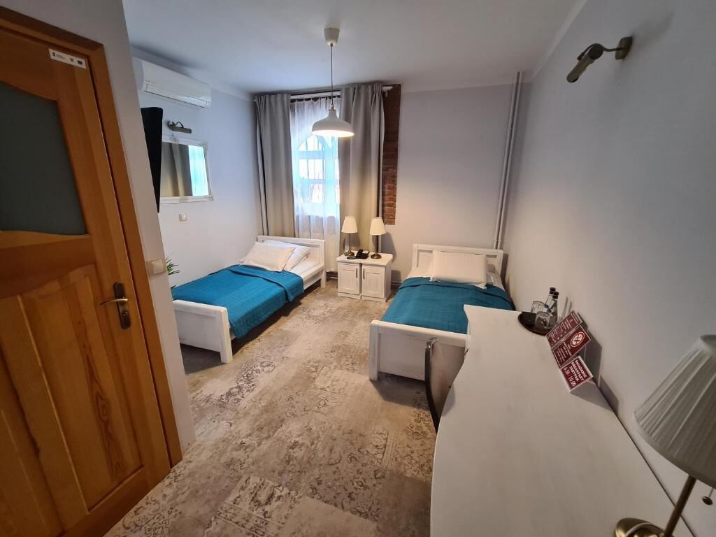Une chambre d’hôtel avec deux lits simples, un bureau, une chaise, une lampe, un radiateur et une porte.