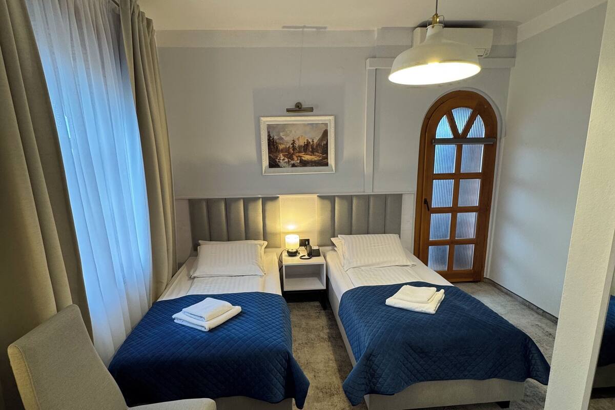 Une chambre d’hôtel avec deux lits, un bureau, une chaise, une table de chevet, une lampe, un tableau au mur et une fenêtre avec des rideaux.