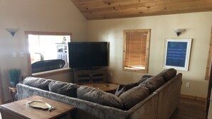 Ett vardagsrum med en stor grå hörnsoffa, ett soffbord i trä, en tv på ett tv-bord och ett fönster med persienner.