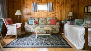 Ein gemütliches Blockhaus-Interieur mit einer blumigen Couch, einem Holztisch, einem Korbstuhl und einem Bett mit weißer Bettdecke.