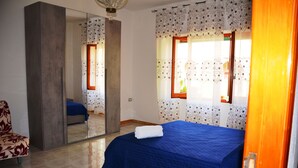 A bedroom with a large mirror, a bed with a blue bedspread, a chair with a floral pattern, and patterned curtains. - B&b Aloysia - Regalati una Vacanza!!!! (Iglesias)
