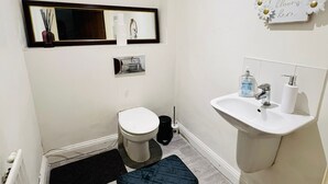 A small bathroom with a toilet, sink, and mirror. There is a black rug on the floor and a white wall with a decorative sign. - Atom Large 4/5 Bed House, Balcony, Sleeps 13, Free Parking, East London (Barking)