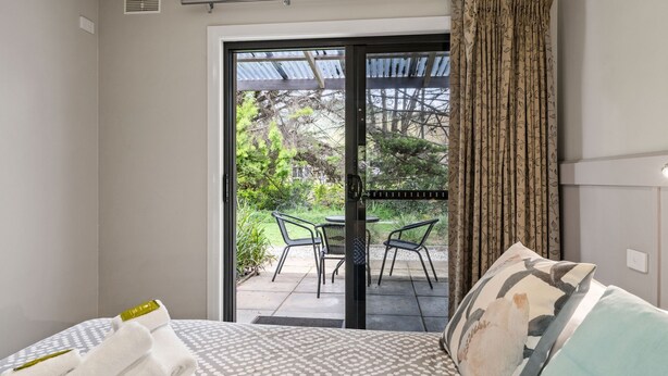 A bedroom with a bed, a window with a view of an outdoor patio, and a curtain. - Grevillea Gardens Unit 5 (BRIGHT)