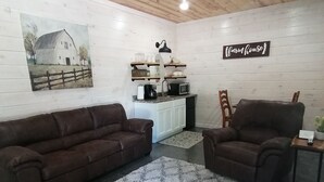 Un salon chaleureux avec un plafond en bois, un ventilateur de plafond, un tableau représentant une grange, un coin cuisine avec un four à micro-ondes et deux canapés marron.