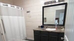 Une salle de bain avec des toilettes blanches, une douche avec un rideau blanc, un lavabo avec un miroir au-dessus, et des meubles en bois foncé.