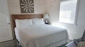 Une chambre avec un plafond en bois, un lit avec une literie blanche, un élément décoratif mural, une lampe de chevet, une fenêtre avec des stores et un tapis à motifs.