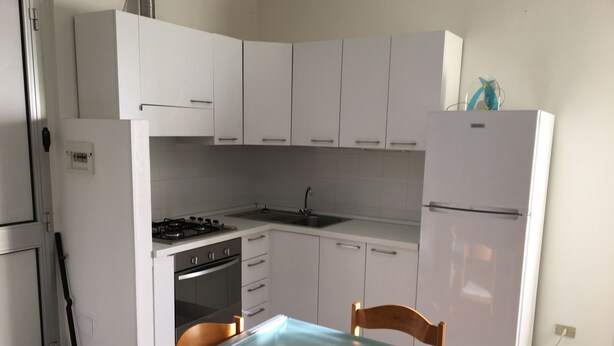 A kitchen with white cabinets, a stove, a refrigerator, and a dining table with chairs. - Sea Fano holiday apartment (Fano)