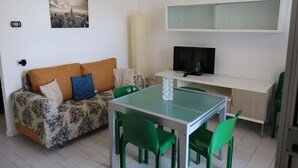 A compact living space with a sofa, a glass-top dining table, and a television. - Sea Fano holiday apartment (Fano)