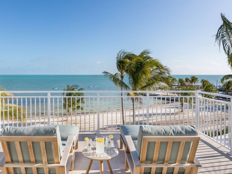 Un balcon avec deux chaises en bois et une petite table, donnant sur une plage bordée de palmiers et sur l’océan.