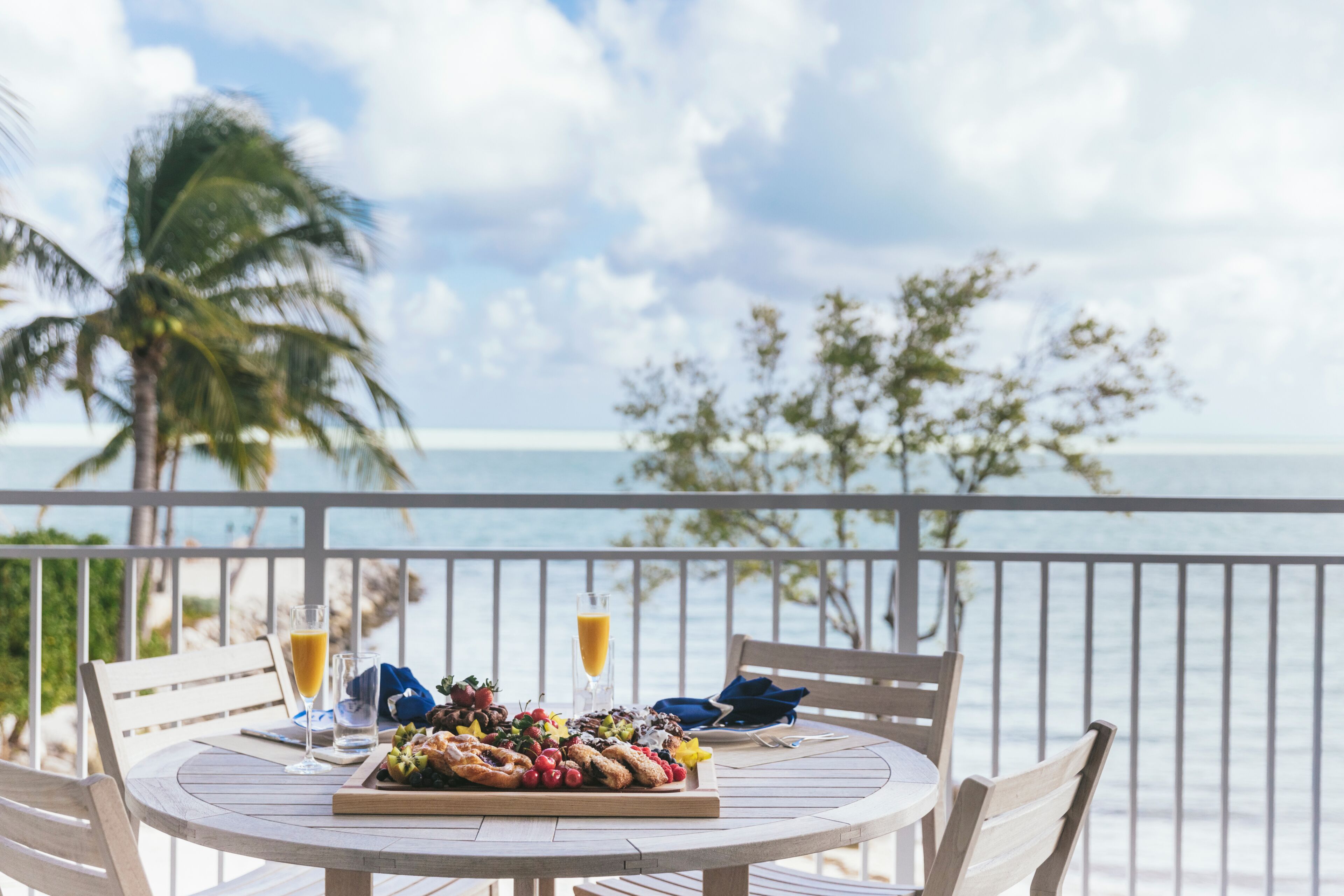 A round wooden table with a fruit platter, two glasses of orange juice, and a blue napkin on a balcony overlooking the sea.