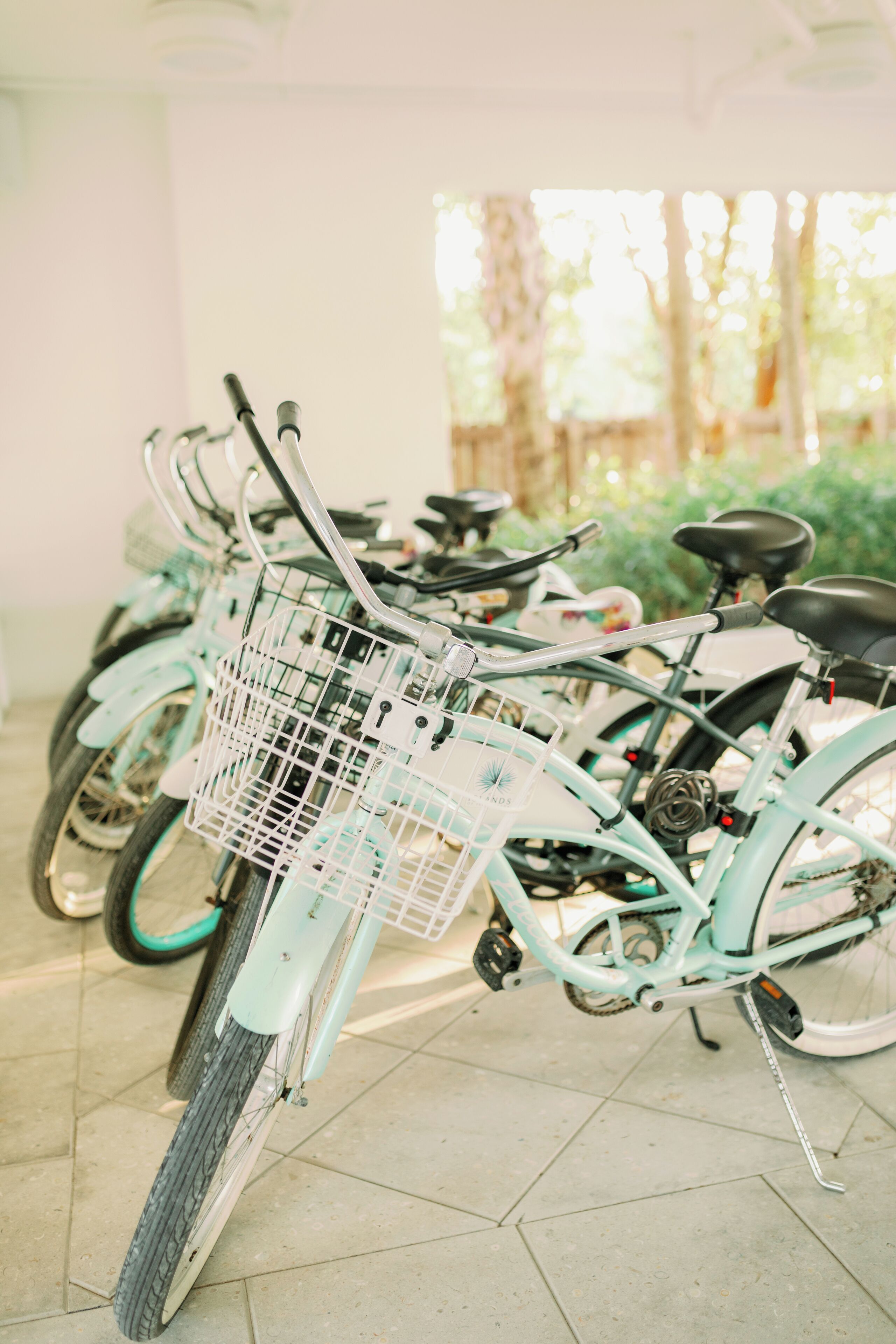 A row of bicycles parked indoors with a tiled floor and a view of greenery outside.