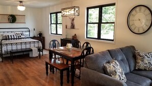 A cozy living room with a sofa, a dining table with chairs, a bed with patterned bedding, and a large wall clock. - Rappahannock River Unplugged (Jamaica)