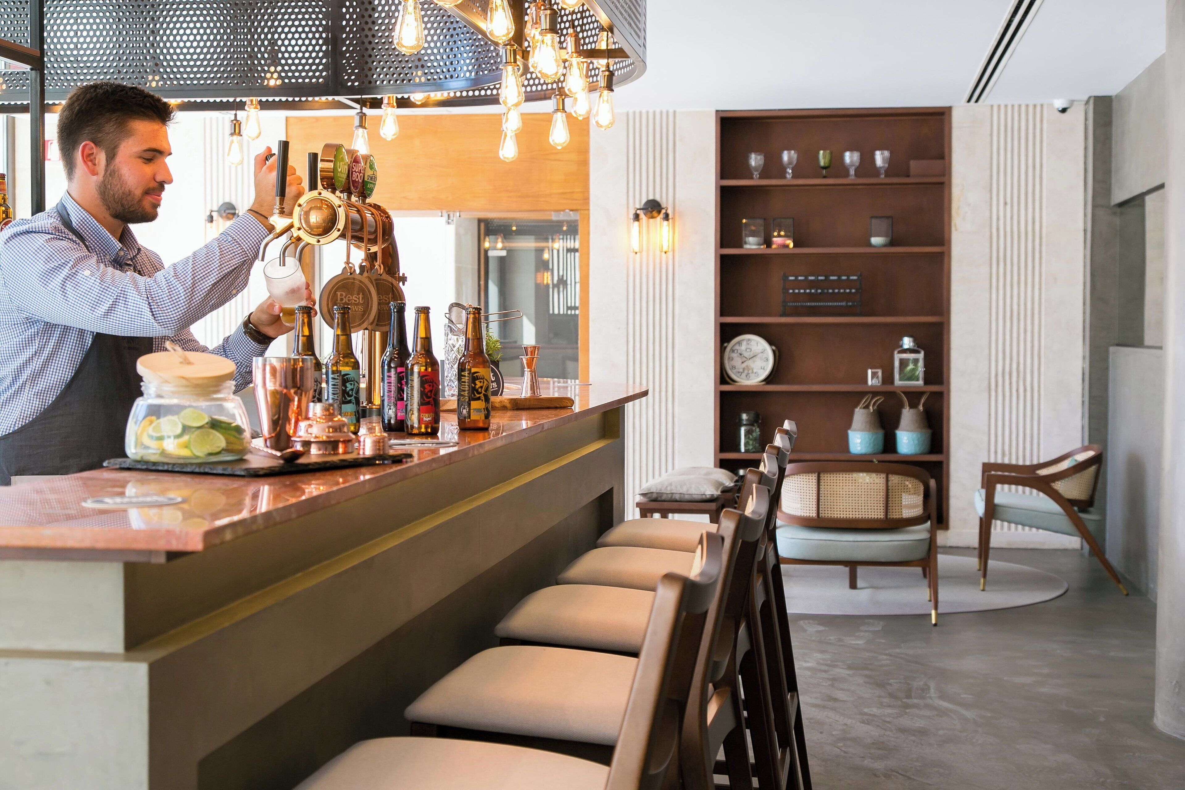 Een bar met een bartender die drankjes maakt, een toonbank met verschillende flessen en een zithoek met stoelen en een ronde tafel.