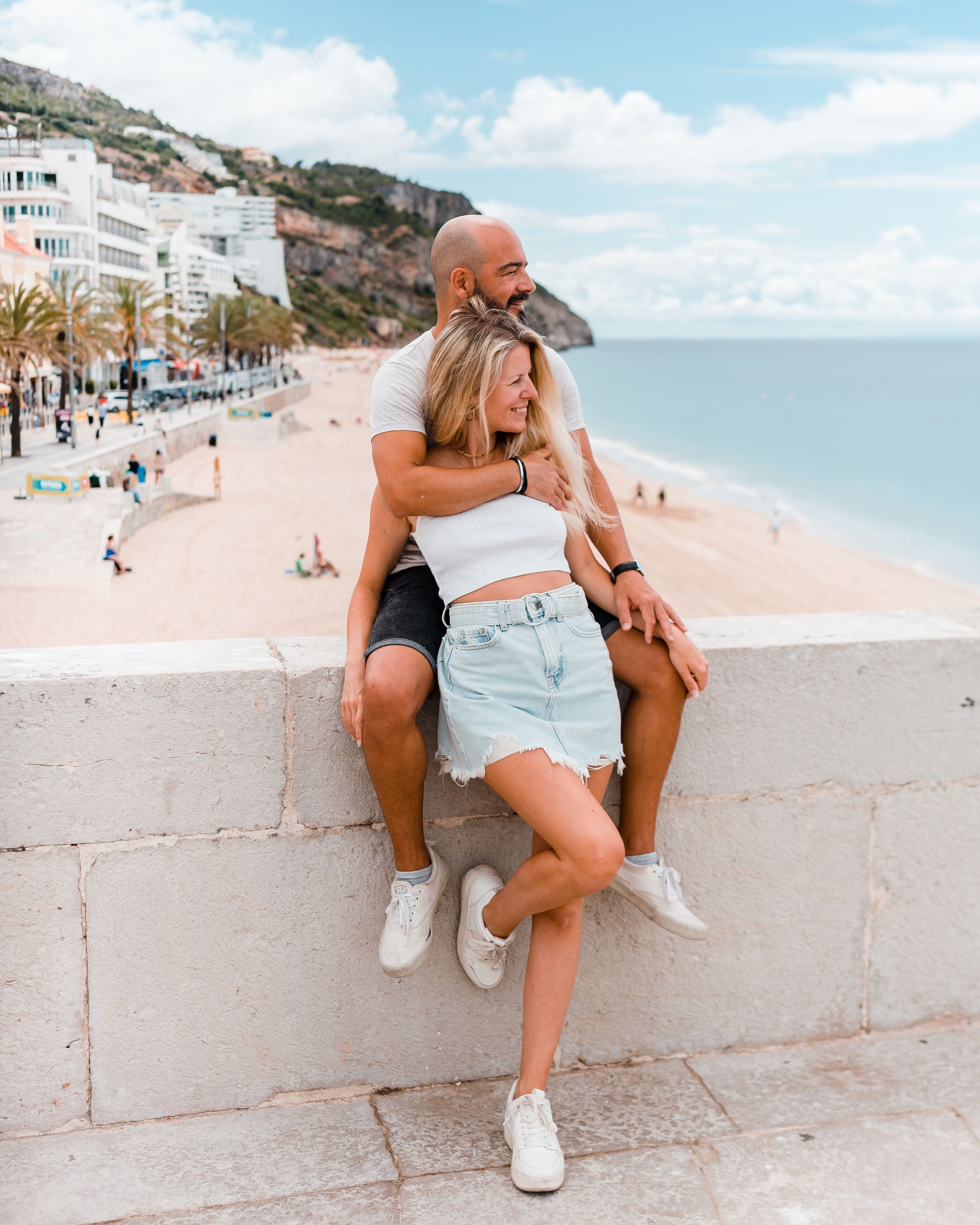 A couple sitting on a stone wall overlooking a beach with buildings and a hilly landscape in the background.