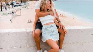A couple sitting on a stone wall overlooking a beach with buildings and a hilly landscape in the background.