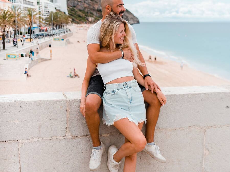 A couple sitting on a stone wall overlooking a beach with buildings and a hilly landscape in the background.