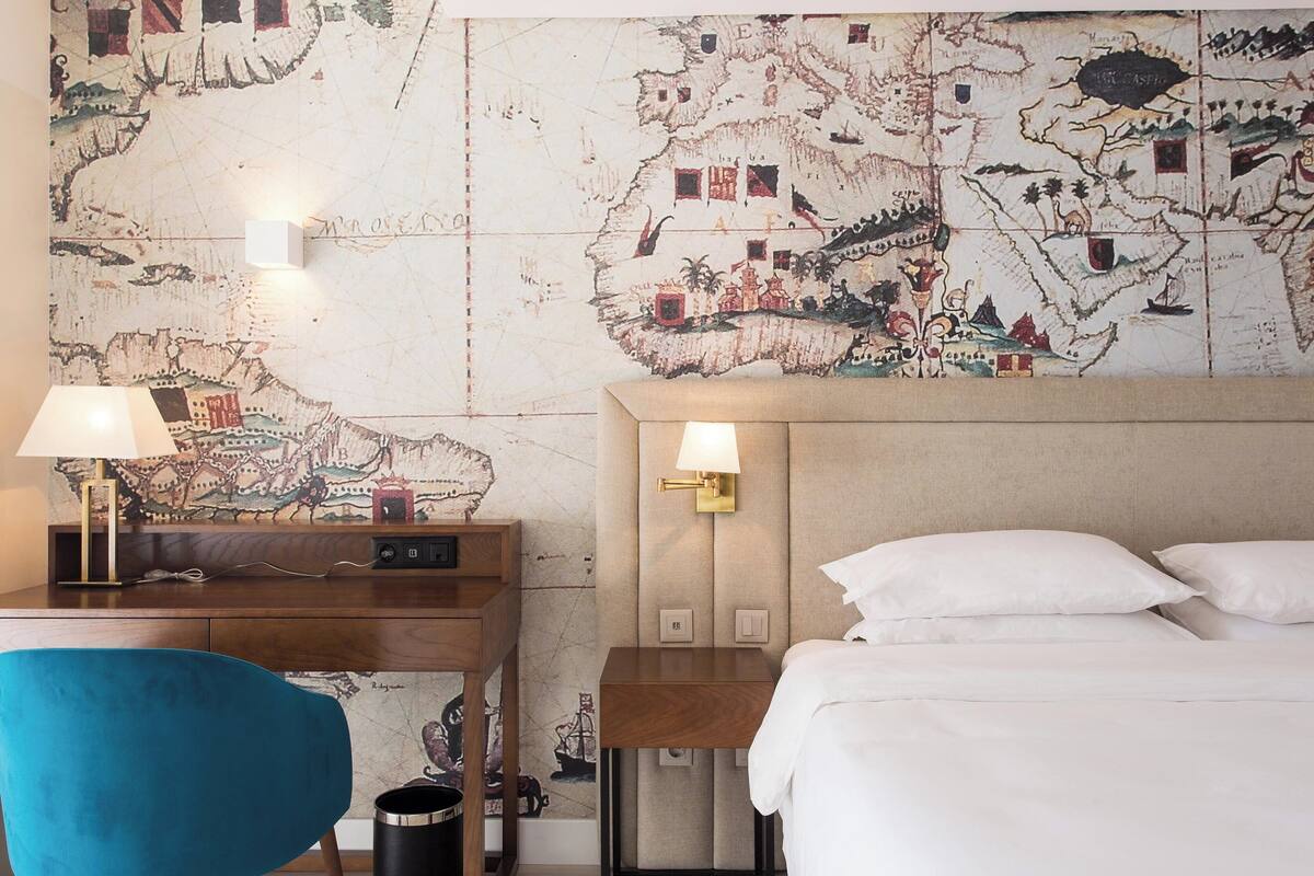 A hotel room with a bed, a desk with a lamp, a chair, and a map on the wall.