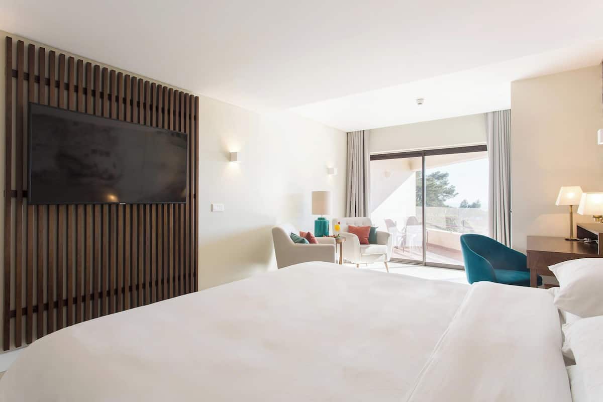 A hotel room with a large bed, a flat-screen TV mounted on a wooden panel, a seating area with chairs, and a balcony with a view.