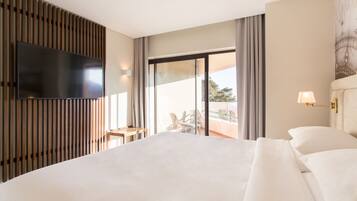 A hotel room with a large bed, a flat-screen TV mounted on a wooden panel, a small table with chairs, and a balcony with a view.