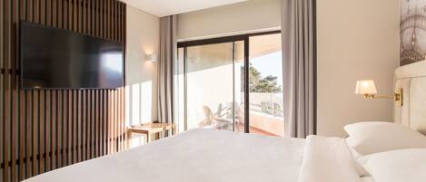 Habitación de hotel con una cama grande, un televisor de pantalla plana montado en un panel de madera, una pequeña mesa con sillas y un balcón con vistas.
