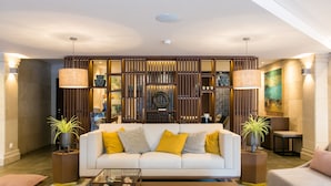 Una sala de estar moderna con un sofá blanco, cojines amarillos y grises, dos mesitas de noche con plantas en macetas y una mesa de centro de vidrio.