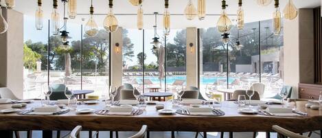 Un comedor con vista a la piscina, con una mesa larga dispuesta para una comida, lámparas colgantes y una decoración interior moderna.