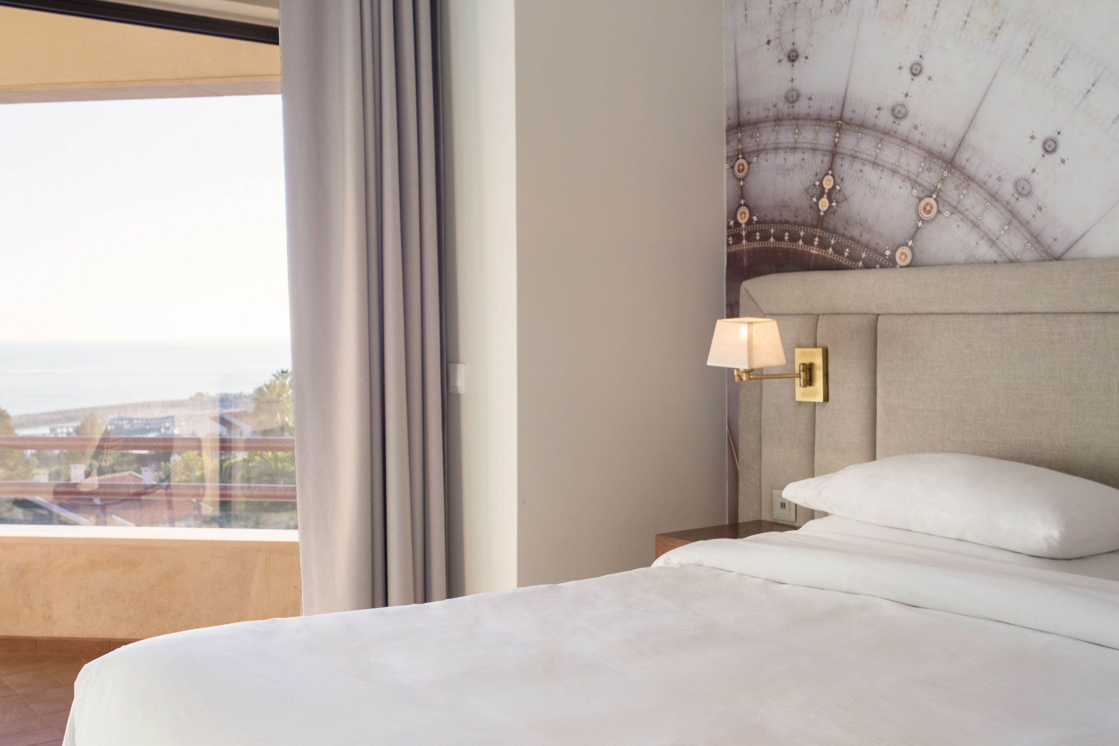 A hotel room with a large bed, a nightstand, a lamp, and a view of the city through the window.